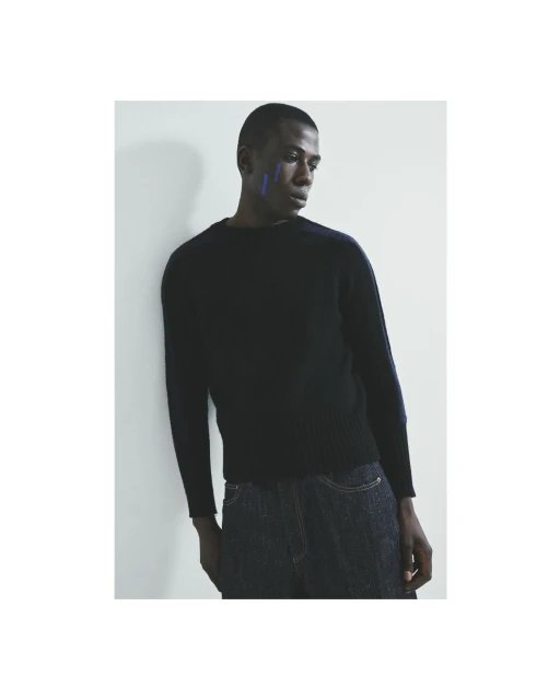 Studio shot of a Black male model in a sweater and jeans, leaning against a white wall, with blue face paint, striking a casual pose.