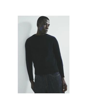 Studio shot of a Black male model in a sweater and jeans, leaning against a white wall, with blue face paint, striking a casual pose.