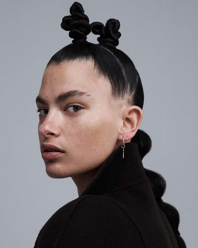 Close-up portrait of a Black female with unique hairstyle, dark jacket, and gold earring set against a neutral grey backdrop.
