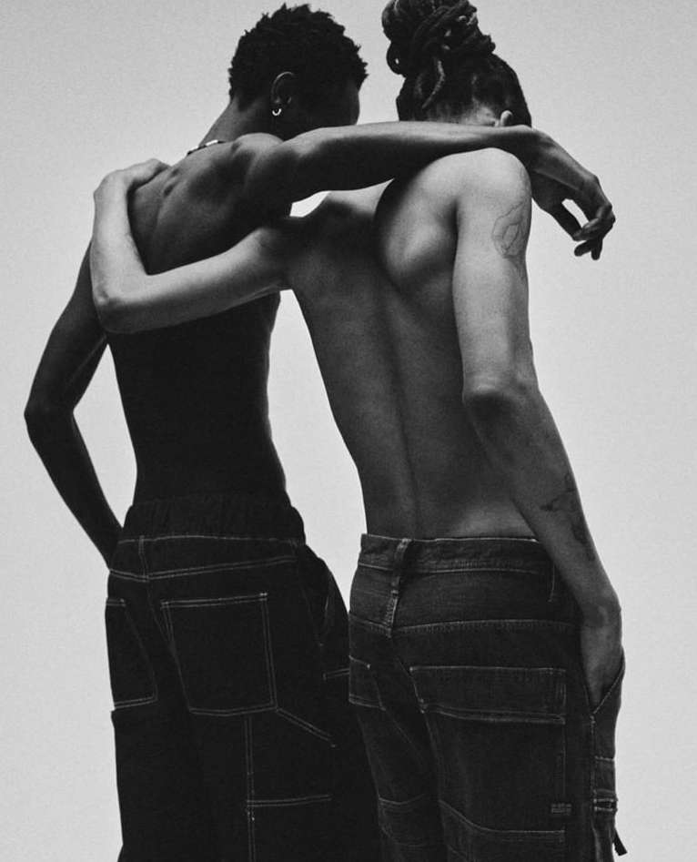 Monochrome portrait of two adult males embracing and wearing denim carpenter pants against a plain background in a photo studio setting.