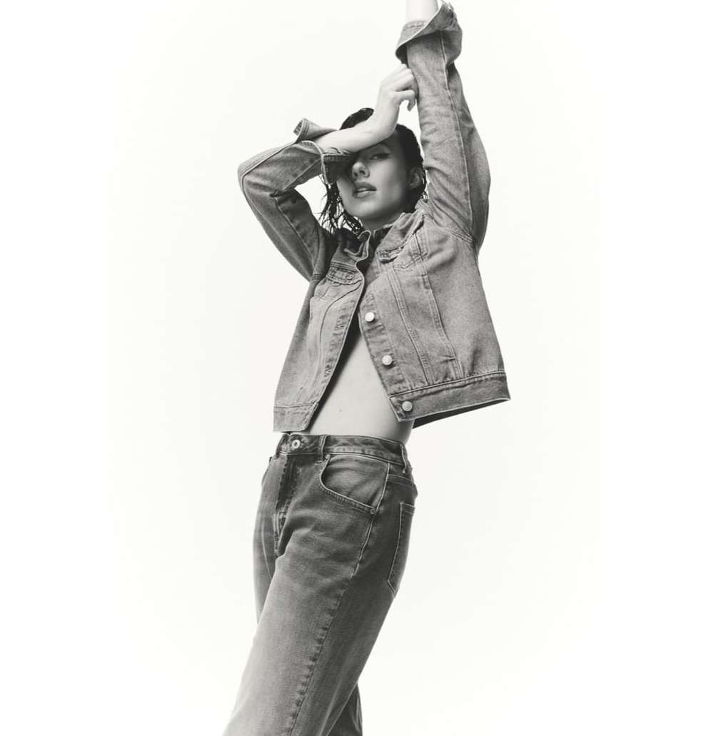 Monochrome fashion shot of a woman in a denim jacket and jeans posing against a white background with her arms raised above her head.