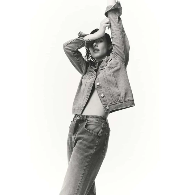 Monochrome fashion shot of a woman in a denim jacket and jeans posing against a white background with her arms raised above her head.