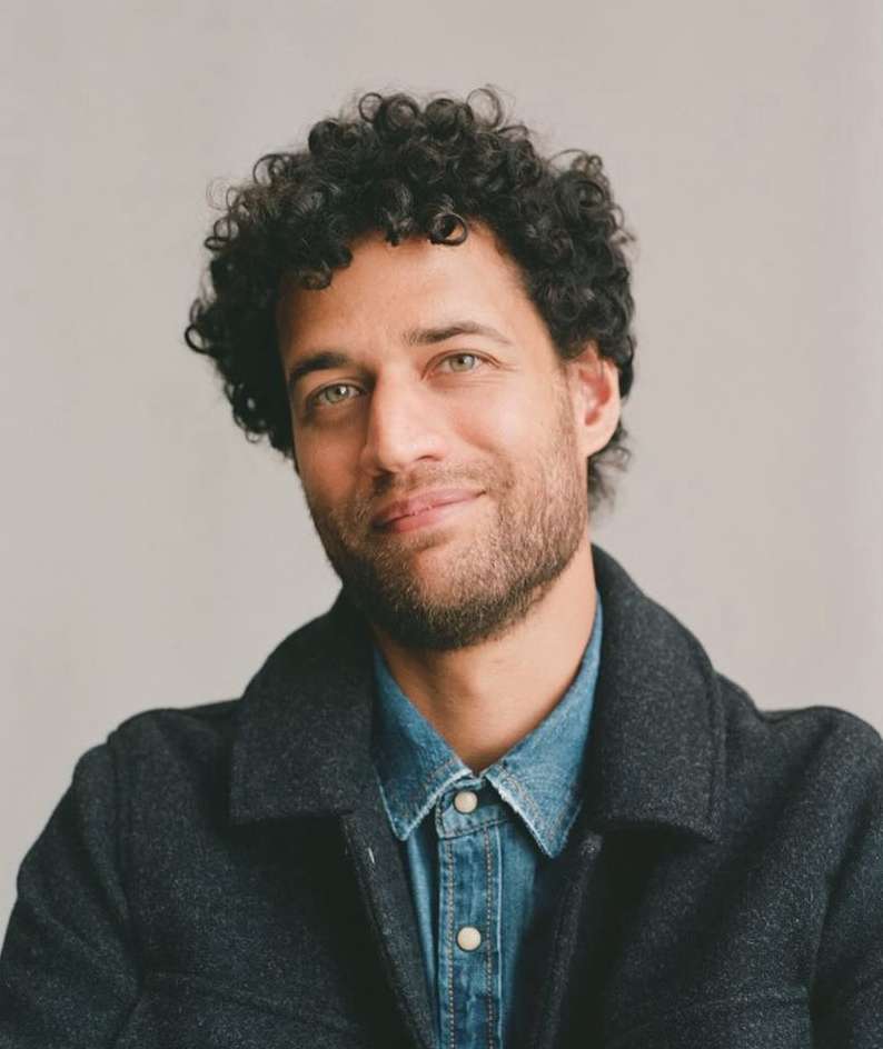 Portrait of a man with curly dark hair and stubble wearing a denim shirt and dark jacket against a plain grey background
