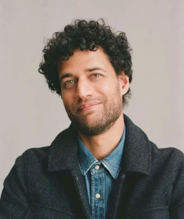 Portrait of a man with curly dark hair and stubble wearing a denim shirt and dark jacket against a plain grey background