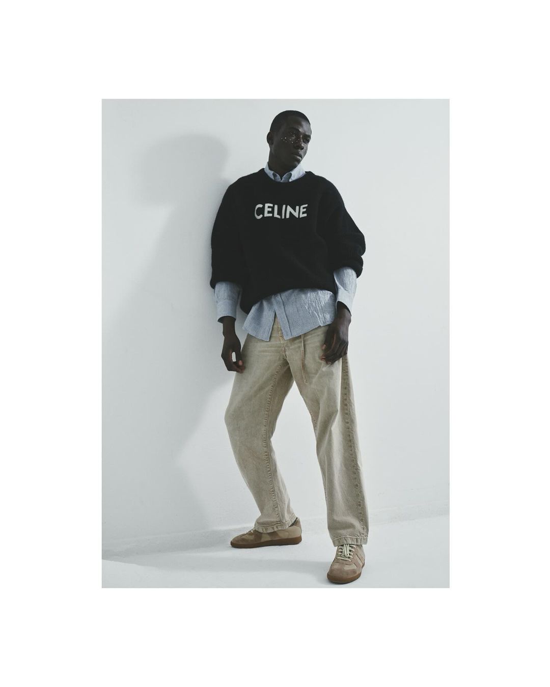 Adult male fashion model in a black CELINE sweater, light blue shirt, beige pants, and brown sneakers in a photo studio setting.