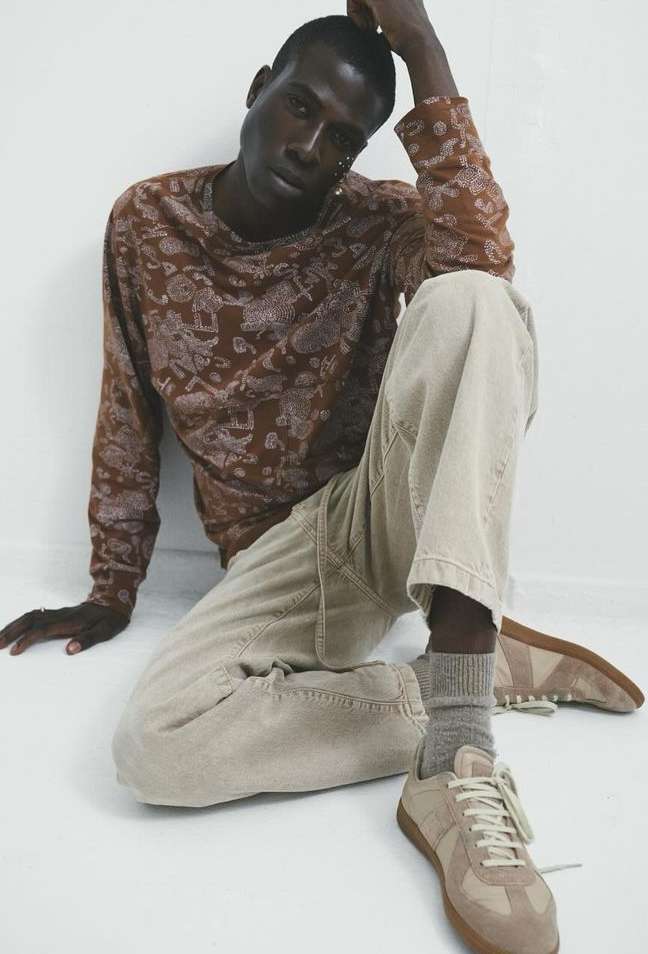 Young man poses in a long-sleeved shirt and pants indoors, seated against a light-colored wall, with a pair of tan shoes.