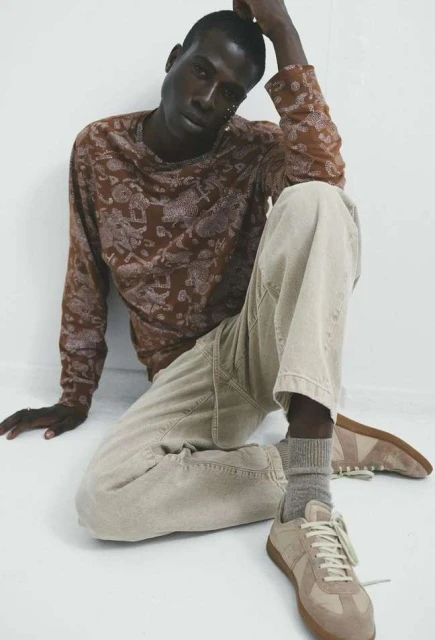 Young man poses in a long-sleeved shirt and pants indoors, seated against a light-colored wall, with a pair of tan shoes.