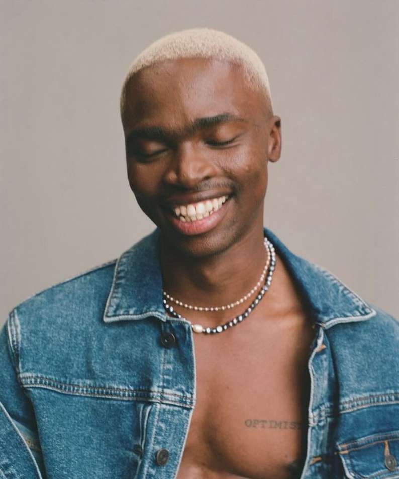 Portrait of smiling Black man with fair hair wearing a denim jacket and necklaces in front of a light-grey neutral backdrop.