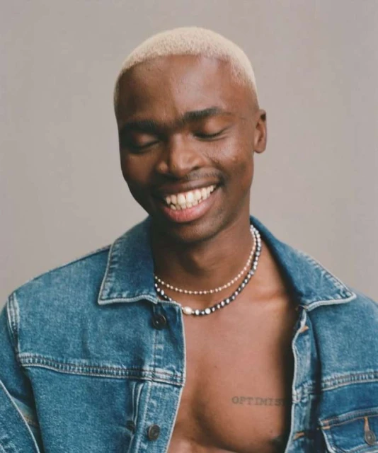 Portrait of smiling Black man with fair hair wearing a denim jacket and necklaces in front of a light-grey neutral backdrop.