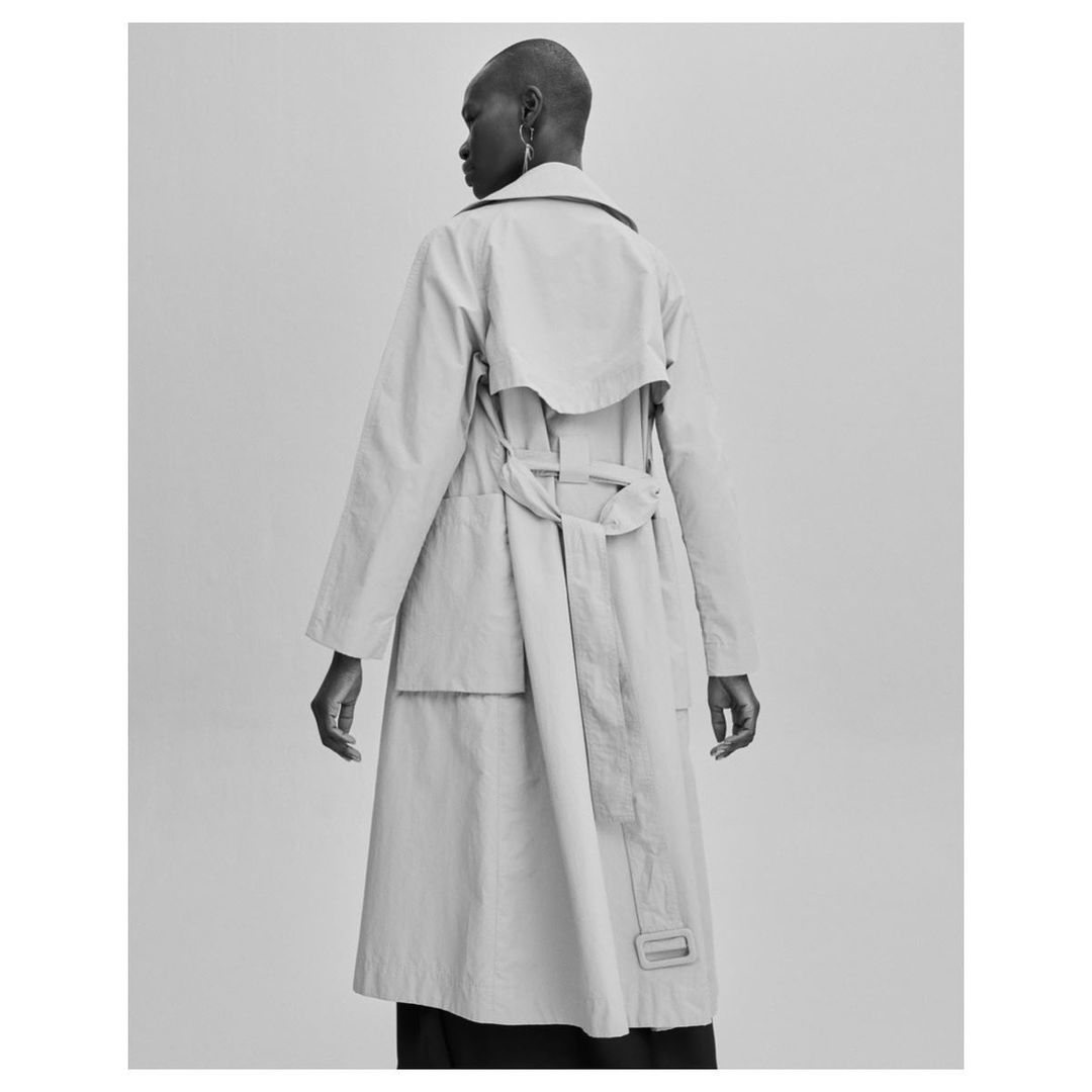 A stylish adult woman with short hair stands in a light studio wearing a beige trench coat, seen from the back.