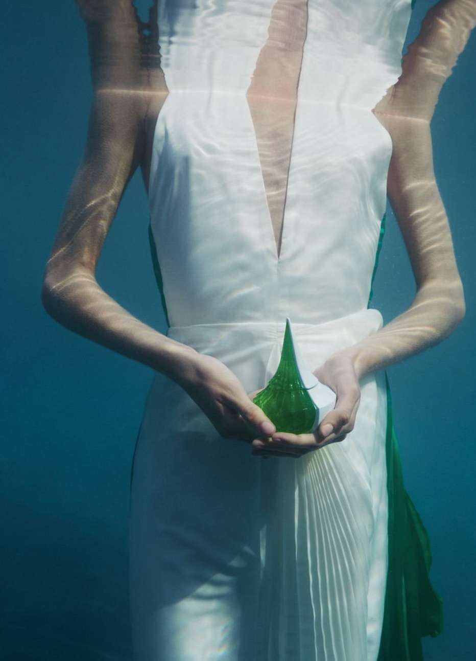 Underwater shot of a woman in a white dress holding a geometric green glass sculpture, creating a surreal and ethereal visual.