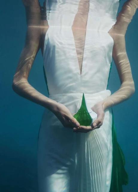 Underwater shot of a woman in a white dress holding a geometric green glass sculpture, creating a surreal and ethereal visual.