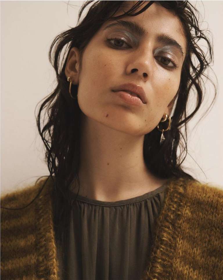 Close-up portrait of a female model with dark hair wearing silver eyeshadow, hoop earrings, and a tan and brown cardigan.
