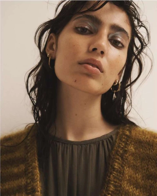 Close-up portrait of a female model with dark hair wearing silver eyeshadow, hoop earrings, and a tan and brown cardigan.