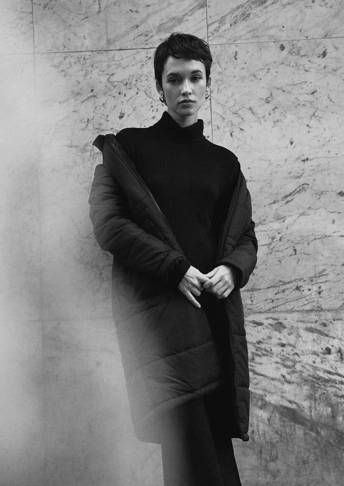 Portrait of a woman with short dark hair wearing a black turtleneck and puffer coat against a marble wall