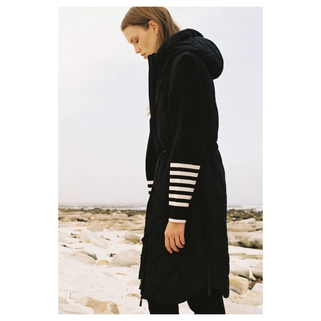 Woman in a black hooded puffer coat with white stripes on the sleeves stands on a beach with a rocky shoreline under an overcast sky.