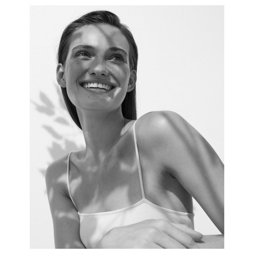 Close-up black and white portrait of a smiling woman with sunlight and shadow patterns on her face and shoulders against a white background.
