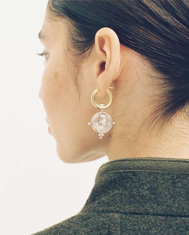 Profile view of an adult woman with dark hair wearing circular gold and decorative silver earrings with white background and green coat.