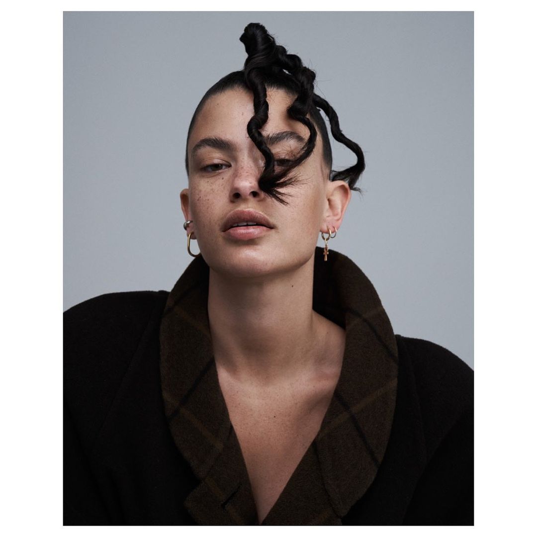 Portrait of an adult female with a unique curly hairstyle and wearing a dark brown blazer in a photo studio with a neutral background.