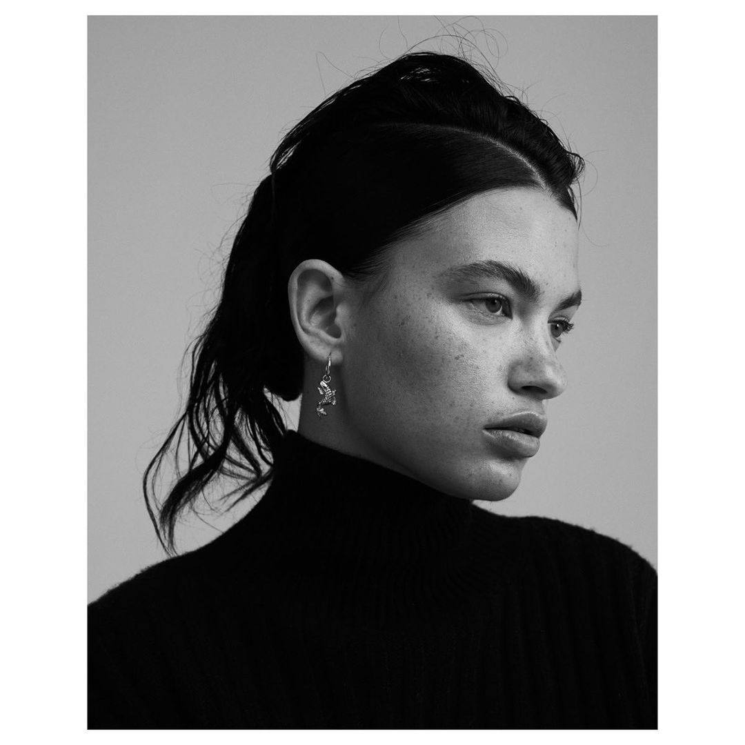 Close-up portrait of a young adult female with light skin, wearing a dark turtleneck, and silver earrings, set against a light background.