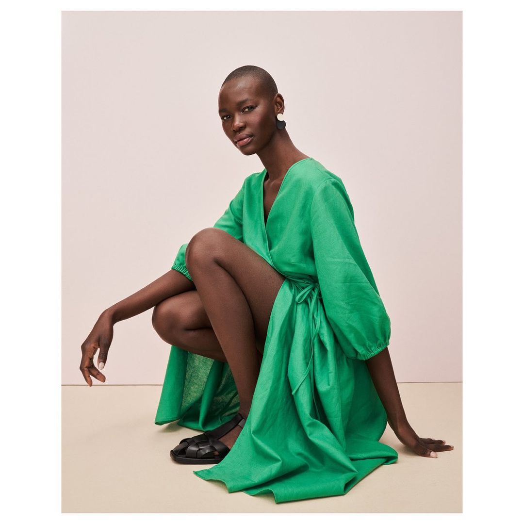 Woman in green wrap dress poses in a photo studio. She is squatting in black sandals against a neutral background with soft lighting.