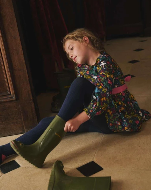 A young girl with blond hair sits on a tiled floor putting on a green rain boot, with another boot beside her, and a floral dress.