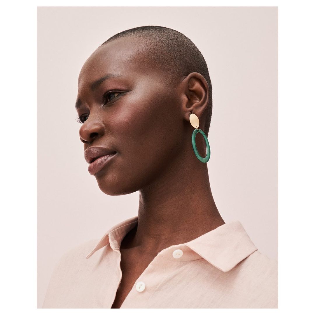 Close-up shot of a stylish adult woman with short hair, wearing an elegant earring, against a clean, light pink background.