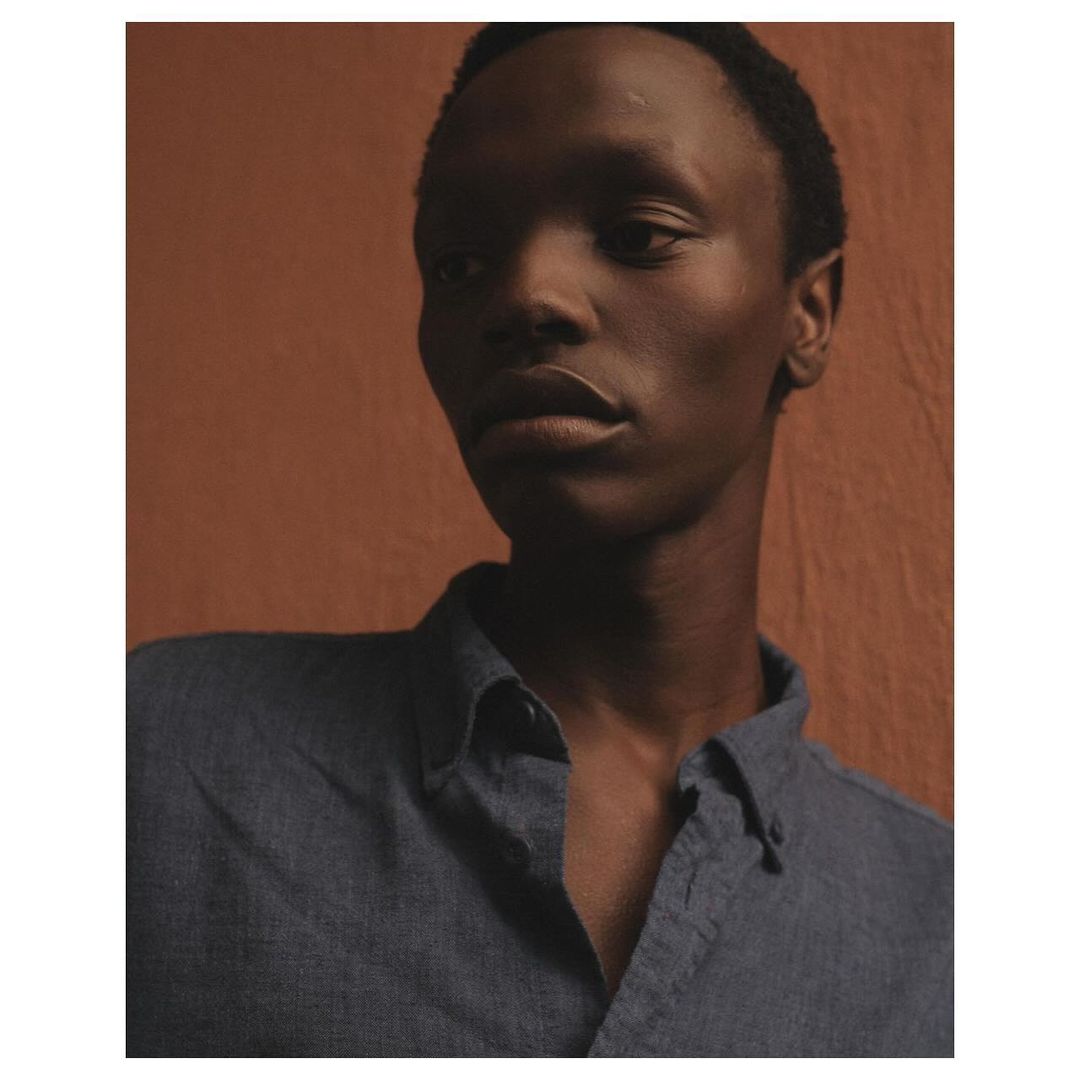 Portrait of a dark-skinned man wearing a blue button-down shirt against a textured terracotta background. He is gazing to the side.