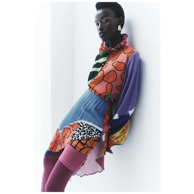 A Black woman poses indoors, wearing a patterned mini dress and pink tights. The garment features long sleeves and a ruffled collar.