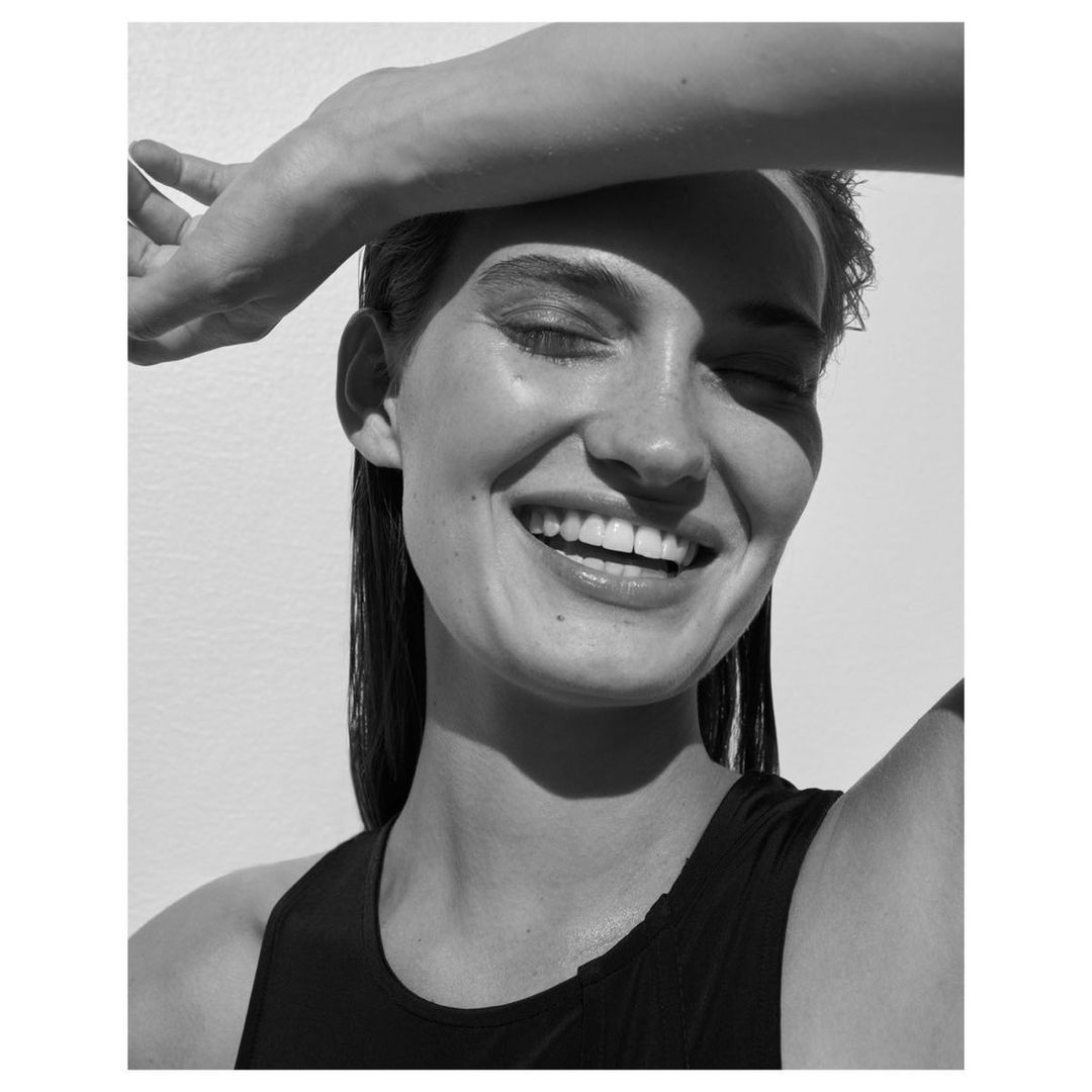 Black and white portrait of a smiling woman with wet hair, eyes closed, and her hand shielding her face from bright sunlight.