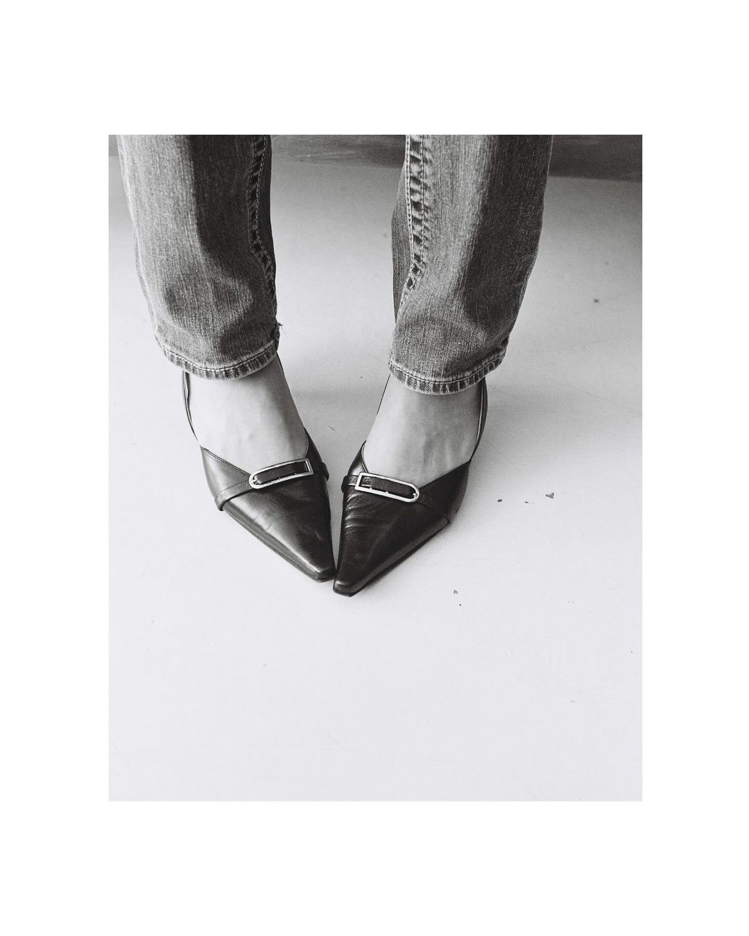 Black and white shot of a woman in blue jeans wearing black pointed-toe high heel shoes, standing on a white surface.