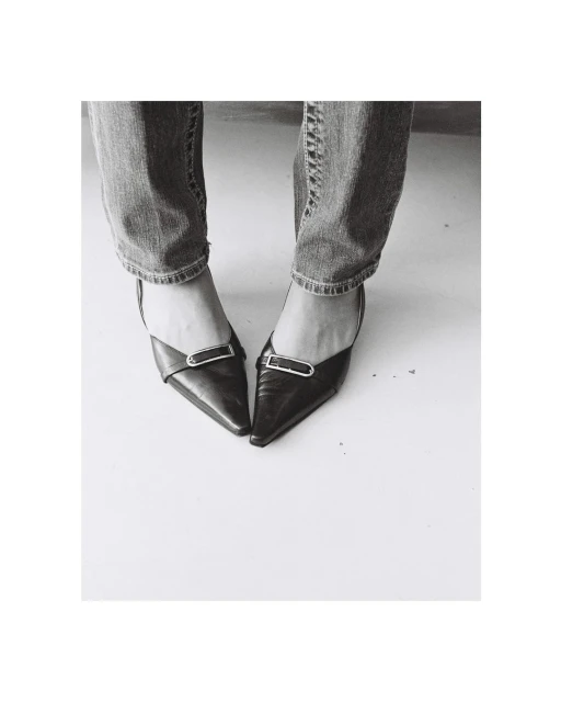 Black and white shot of a woman in blue jeans wearing black pointed-toe high heel shoes, standing on a white surface.