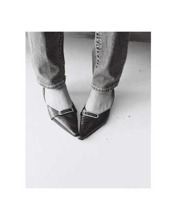 Black and white shot of a woman in blue jeans wearing black pointed-toe high heel shoes, standing on a white surface.
