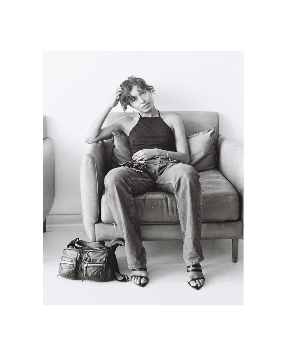 Monochrome portrait of a young woman in a black halter top and jeans, seated in a chair next to a bag, in a minimalistic indoor setting.