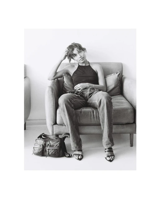 Monochrome portrait of a young woman in a black halter top and jeans, seated in a chair next to a bag, in a minimalistic indoor setting.