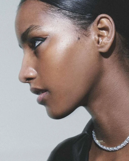 Close-up portrait of a young adult woman with dark hair and brown skin, wearing a black top and a diamond tennis necklace, side profile.
