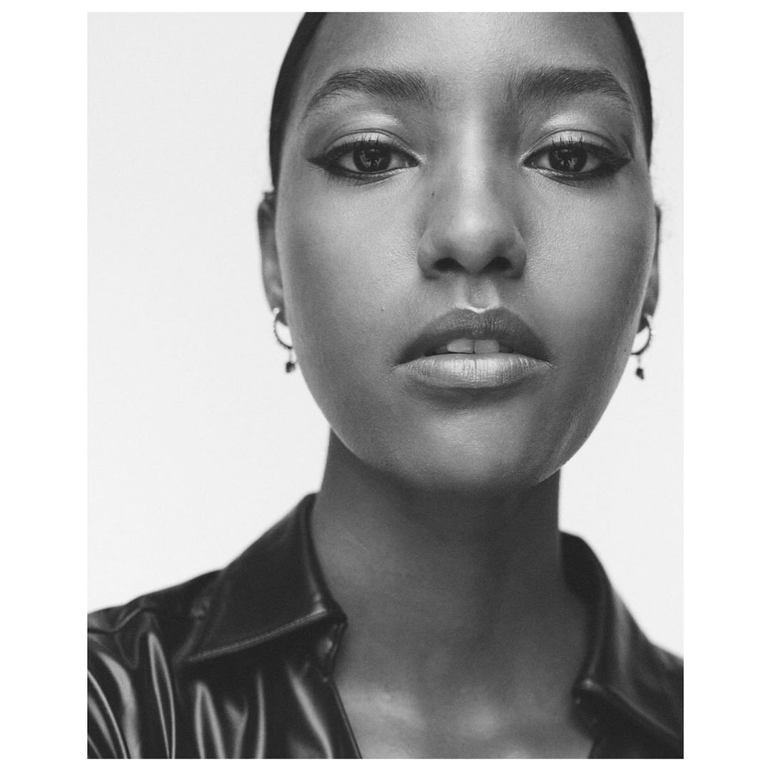 Close-up monochrome portrait of a woman wearing hoop earrings and a dark collar against a plain white background.