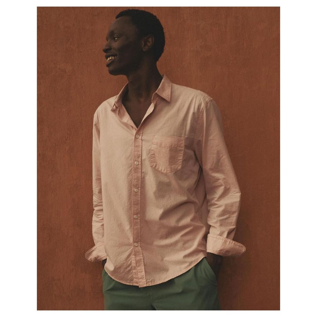 Dark-skinned male model wearing a pink button-down shirt and green trousers poses against a textured orange studio backdrop, smiling.