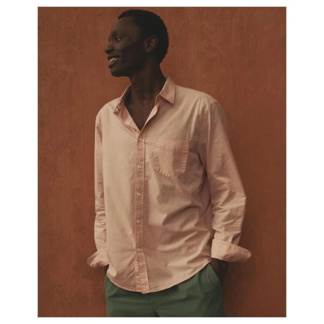Dark-skinned male model wearing a pink button-down shirt and green trousers poses against a textured orange studio backdrop, smiling.