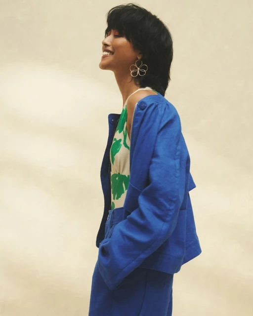 A woman with short black hair smiles in a side profile wearing a blue jacket over a floral print top against a tan backdrop.