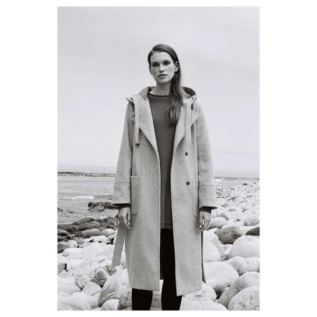 Woman in a hooded grey coat stands on a rocky beach against a pale sky; she is the main focus of the black and white fashion shot.