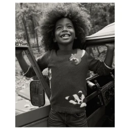 A smiling child with voluminous curly hair wearing a patterned knit sweater leaning against a vintage vehicle in a woodland setting
