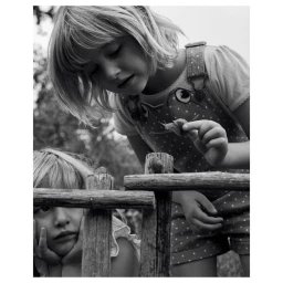 Two young children outdoors near a wooden fence, with one child holding a small snail.