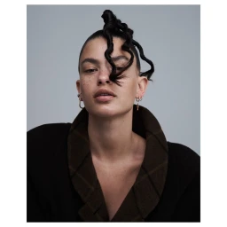 Portrait of a woman with sculpted dark hair and hoop earrings wearing a dark plaid collar against a grey background