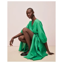 Black woman wearing a long green wrap dress and black sandals, crouching against a pale neutral studio backdrop.