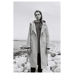 Monochrome portrait of a woman wearing a long belted coat standing on a rocky beach by the ocean, photographed by Luke Kuisis.