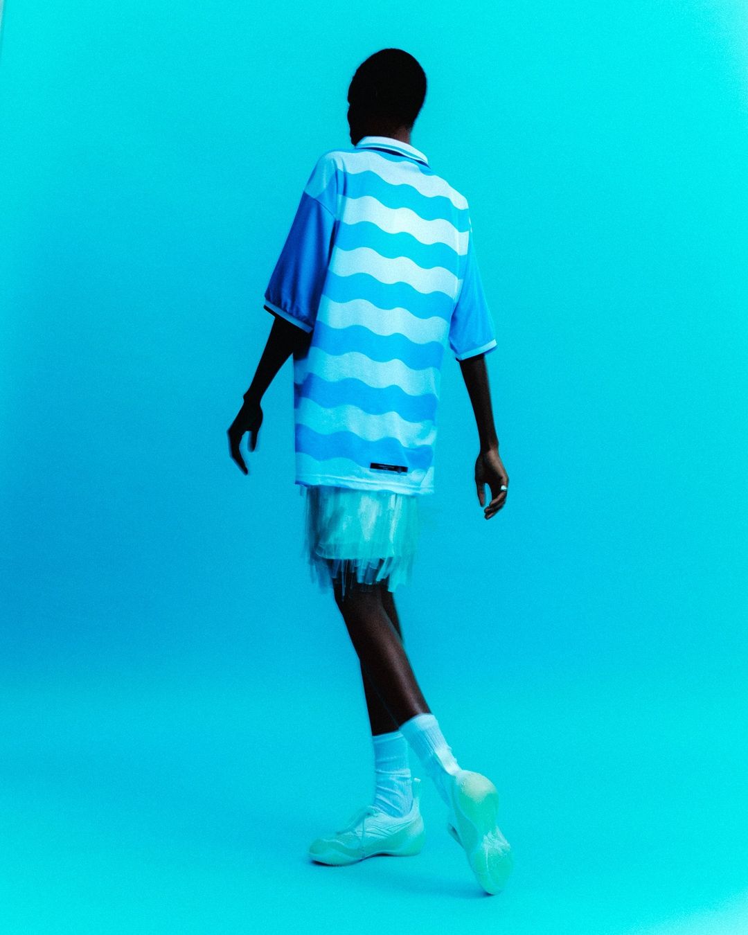 Back view of a Black male model in blue wave shirt, white fringed shorts, socks, and sneakers against a solid bright blue background.