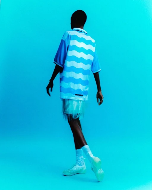 Back view of a Black male model in blue wave shirt, white fringed shorts, socks, and sneakers against a solid bright blue background.