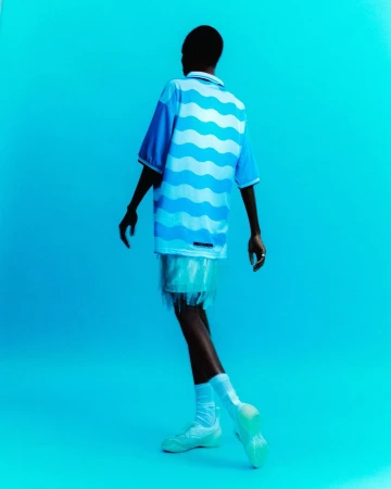 Back view of a Black male model in blue wave shirt, white fringed shorts, socks, and sneakers against a solid bright blue background.