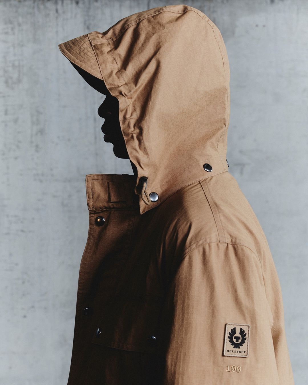 Profile shot of a person wearing a tan hooded jacket with visible button and logo details against a textured gray backdrop in studio lighting.